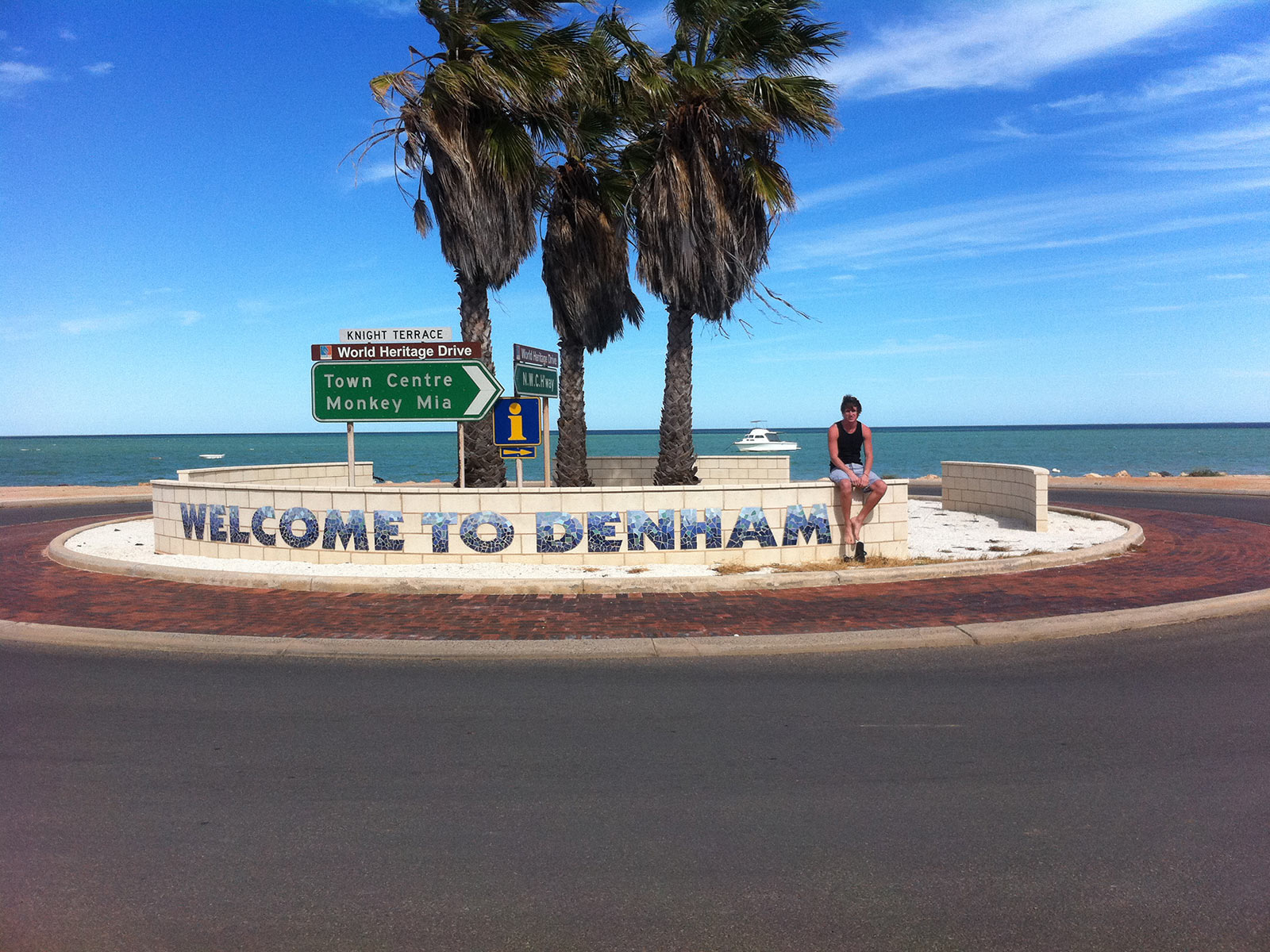David Simpson with Welcome to Denham sign in West Coast, Australia. Hitting a cow on the west coast road trip