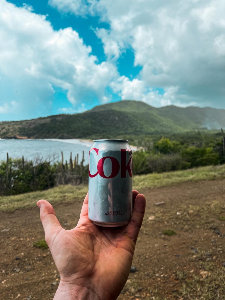 Coke in can on hand with back drop of mountains on a cloudy day in Antigua. Rendezvous beach and cliffside accom in BVI