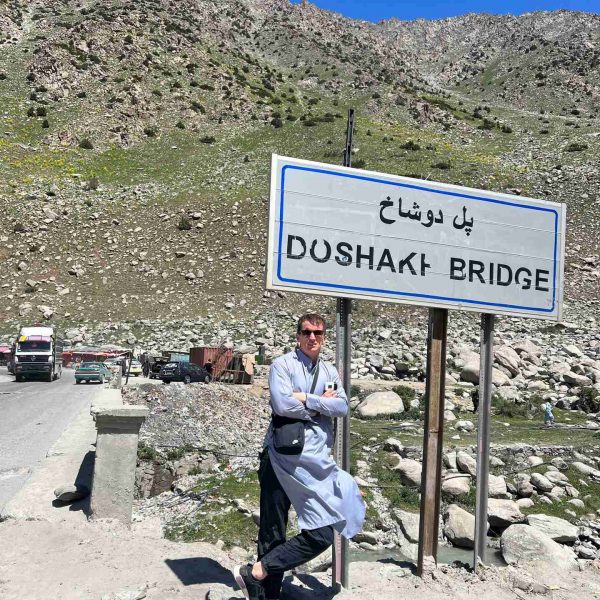 David Simpson standing by the bridge sign in Mazar, Afghanistan. Body search, Soviet exploits & The Salang Pass